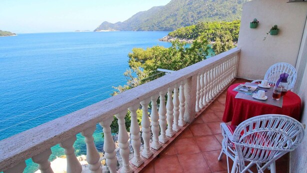 A balcony with a view of the sea and mountains, white wicker chairs, and a red tablecloth.