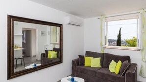 A modern living room with a large mirror, a brown sofa with yellow cushions, a small white table, and a window with a view of the city. - Apartment Semi - One Bedroom Apartment With Terrace and Sea View (Dubrovnik)
