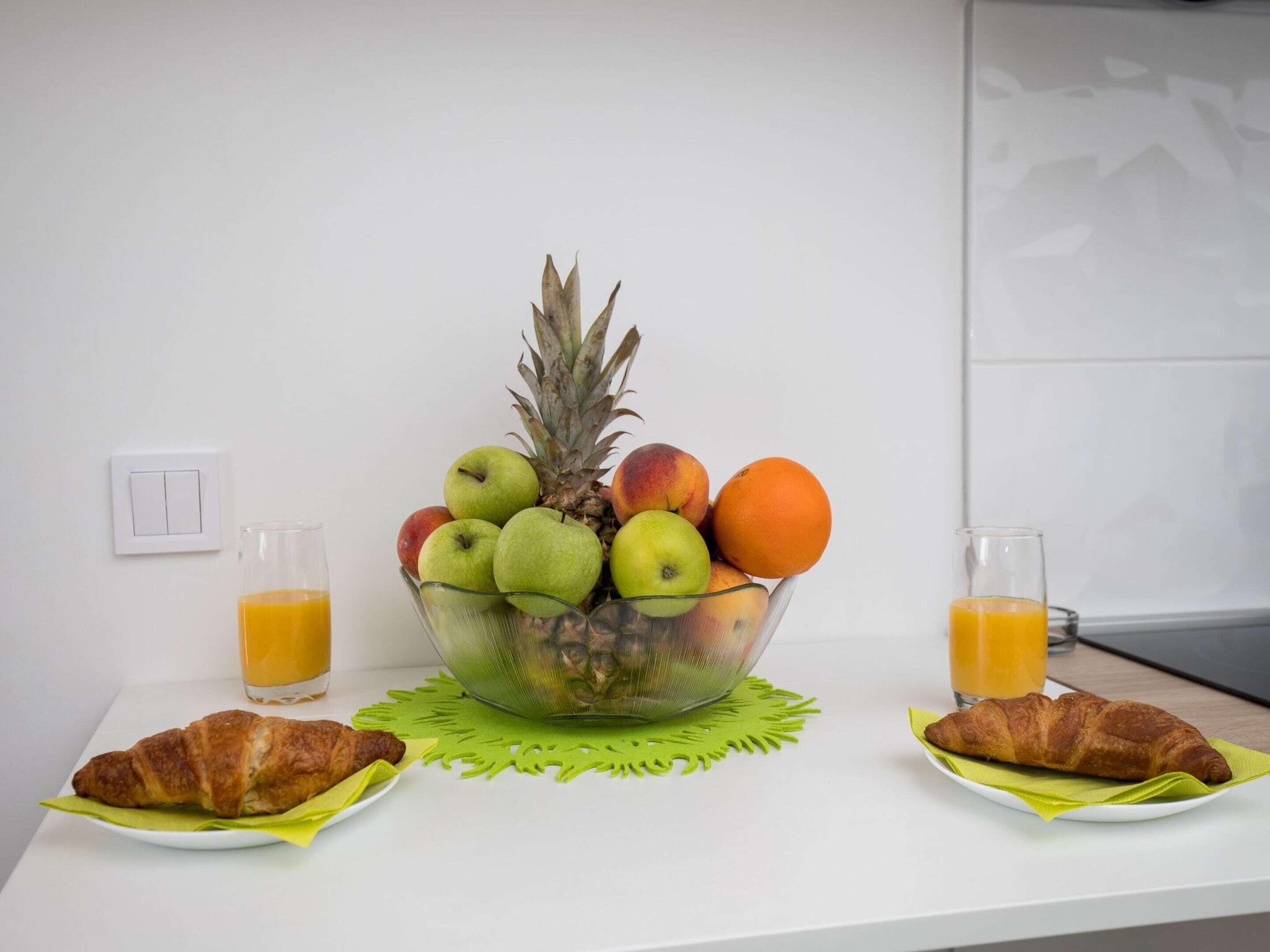 Uma mesa branca com frutas, croissants e copos de suco de laranja.