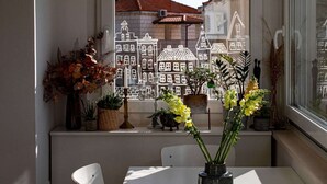 Une vue à travers une fenêtre, avec une silhouette de bâtiments découpée dans du papier blanc, une table avec des chaises et divers plantes en pot.