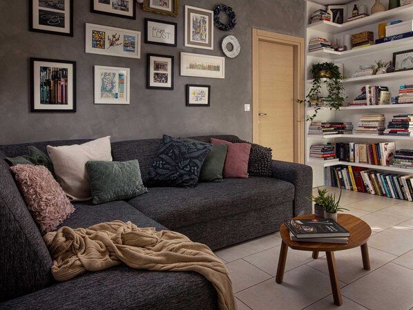 Un salon avec un canapé gris, de nombreux coussins, une table basse sur laquelle repose un livre, et une bibliothèque remplie de livres.