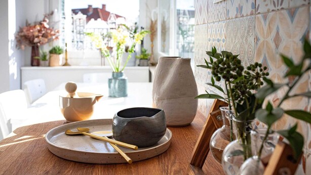 Une table en bois sur laquelle se trouvent un bol en céramique, une cuillère et une théière. On aperçoit, à l’arrière-plan, des plantes en pot et un mur recouvert de carreaux.