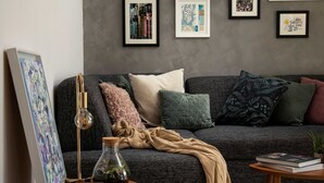 Un salon chaleureux avec un canapé gris, divers coussins, une petite table en bois sur laquelle sont posés un vase et un bol, et des œuvres d’art encadrées accrochées au mur.