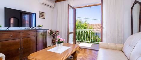 Una habitación con una mesa de madera, un sofá, un televisor, un armario y un balcón con vistas a zonas verdes.