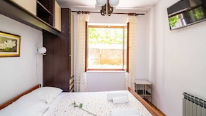Un dormitorio con cama, ventana con cortinas, radiador, una mesita y televisor fijado a la pared.
