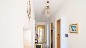 Un pasillo con pared blanca, un banco de madera, una mesita con una computadora portátil, un candelabro colgante y un cuadro en la pared.