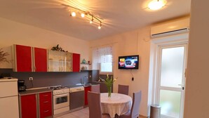 Een kleine keuken met rode kastjes, een fornuis en een spoelbak. Er staat een eettafel met stoelen en een televisie hangt aan de muur.