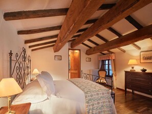 Un dormitorio con techo de madera, una cama con colcha estampada, una mesita de noche con lámpara, un aparador y una ventana con cortinas.