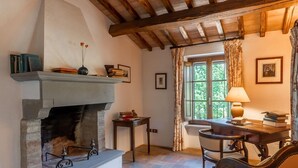 Un cuarto acogedor con chimenea, una mesa con libros, una silla y un ventanal con cortinas.