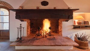 Una chimenea con un revestimiento de piedra, una repisa con tres macetas metálicas y un estante de madera con libros y una lámpara.