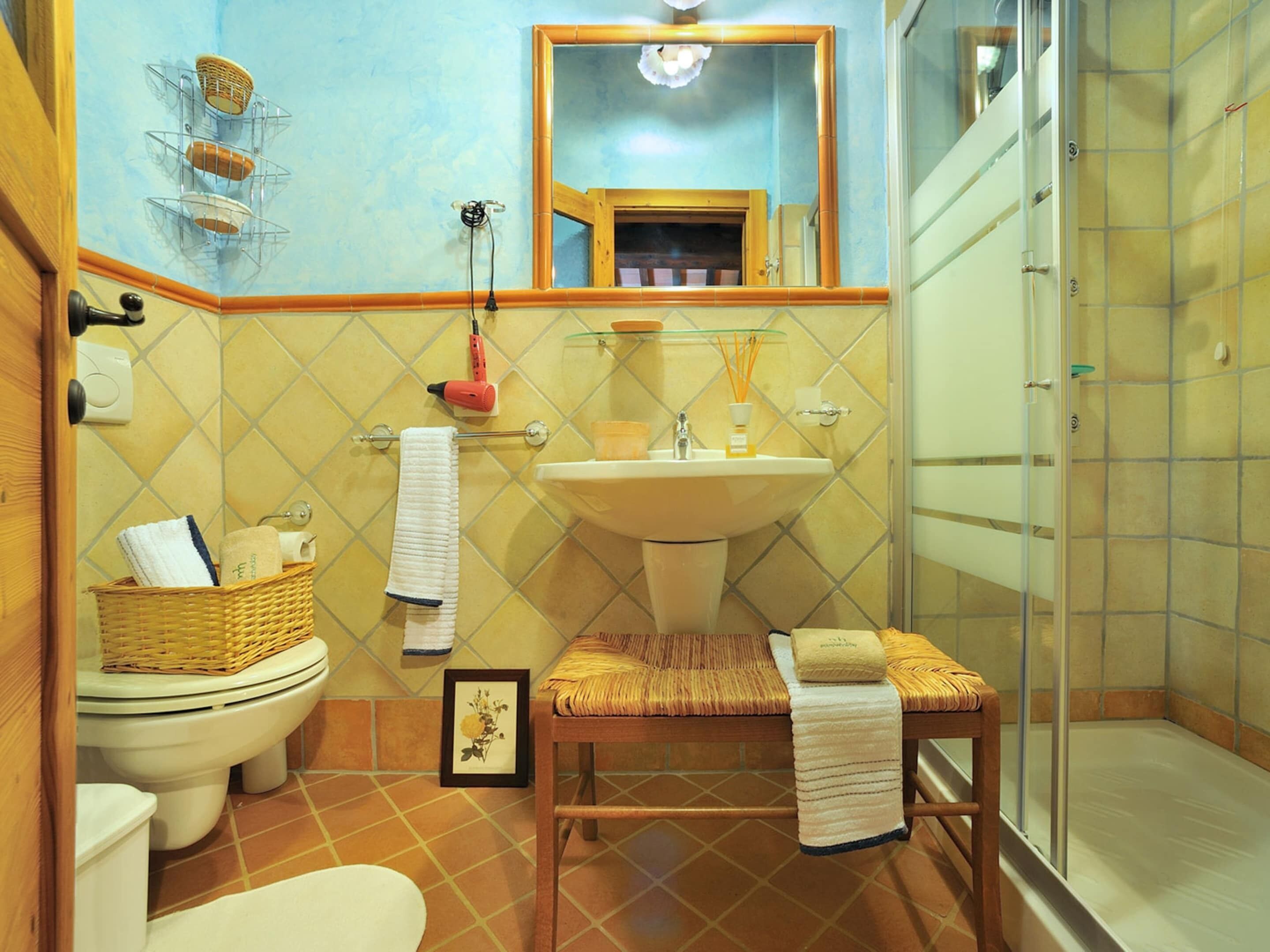 Une salle de bain avec des toilettes, un lavabo et une douche. On y trouve un panier en osier sur les toilettes, un tableau encadré posé sur le sol et une serviette sur la chaise.