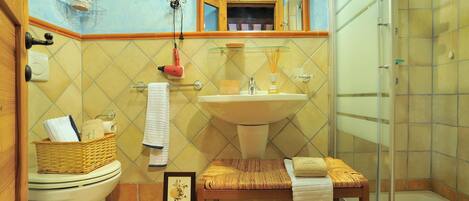 Une salle de bain avec des toilettes, un lavabo et une douche. On y trouve un panier en osier sur les toilettes, un tableau encadré posé sur le sol et une serviette sur la chaise.