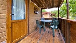 A wooden deck with a table and chairs, a door with a window, and a view of a building in the background. - Belvilla by OYO Lovely Bungalow With a Shared Pool (Capraia e Limite)