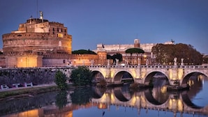 Un ponte storico con archi che si riflettono nell'acqua, un grande edificio simile a una fortezza e una struttura a cupola sullo sfondo.