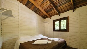 Un letto singolo con lenzuola bianche e copriletto marrone, una finestra vista verde, e travi in legno al soffitto.