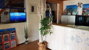 A reception area with a wooden reception desk, a television displaying a scenic view, a wall-mounted life preserver decoration, and a bulletin board with travel posters. - Apartment Near Golfo di Cugnana (Olbia)
