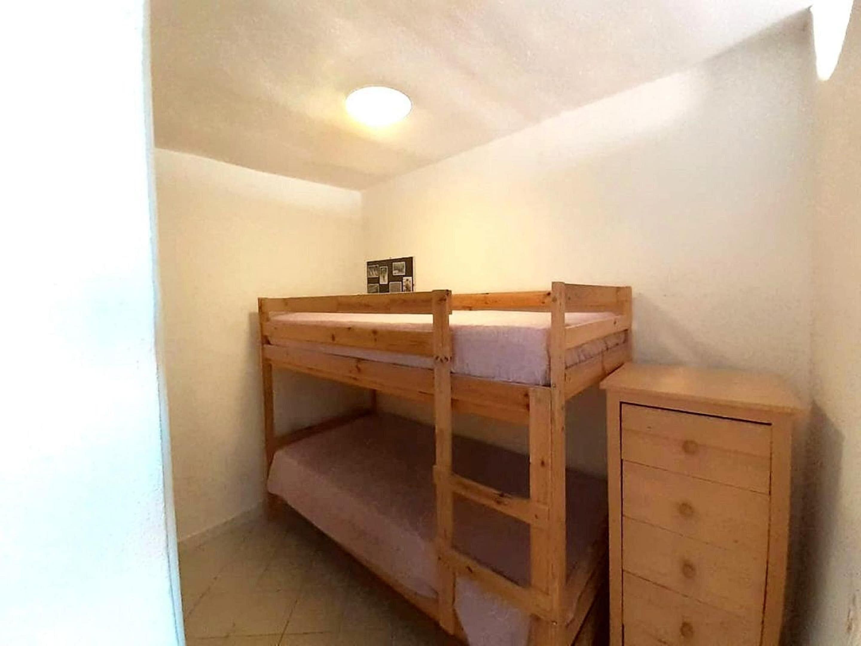 A bunk bed with a lower shelf and a small cabinet, in a room with white walls and a ceiling light.