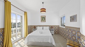 Habitación de hotel con una cama grande, una mesita de noche, una silla, un ventanal y un balcón con cortinas amarillas.
