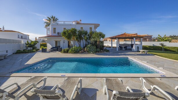 Una piscina con sillas de descanso, un patio cubierto y un edificio blanco con tejado de azulejos.