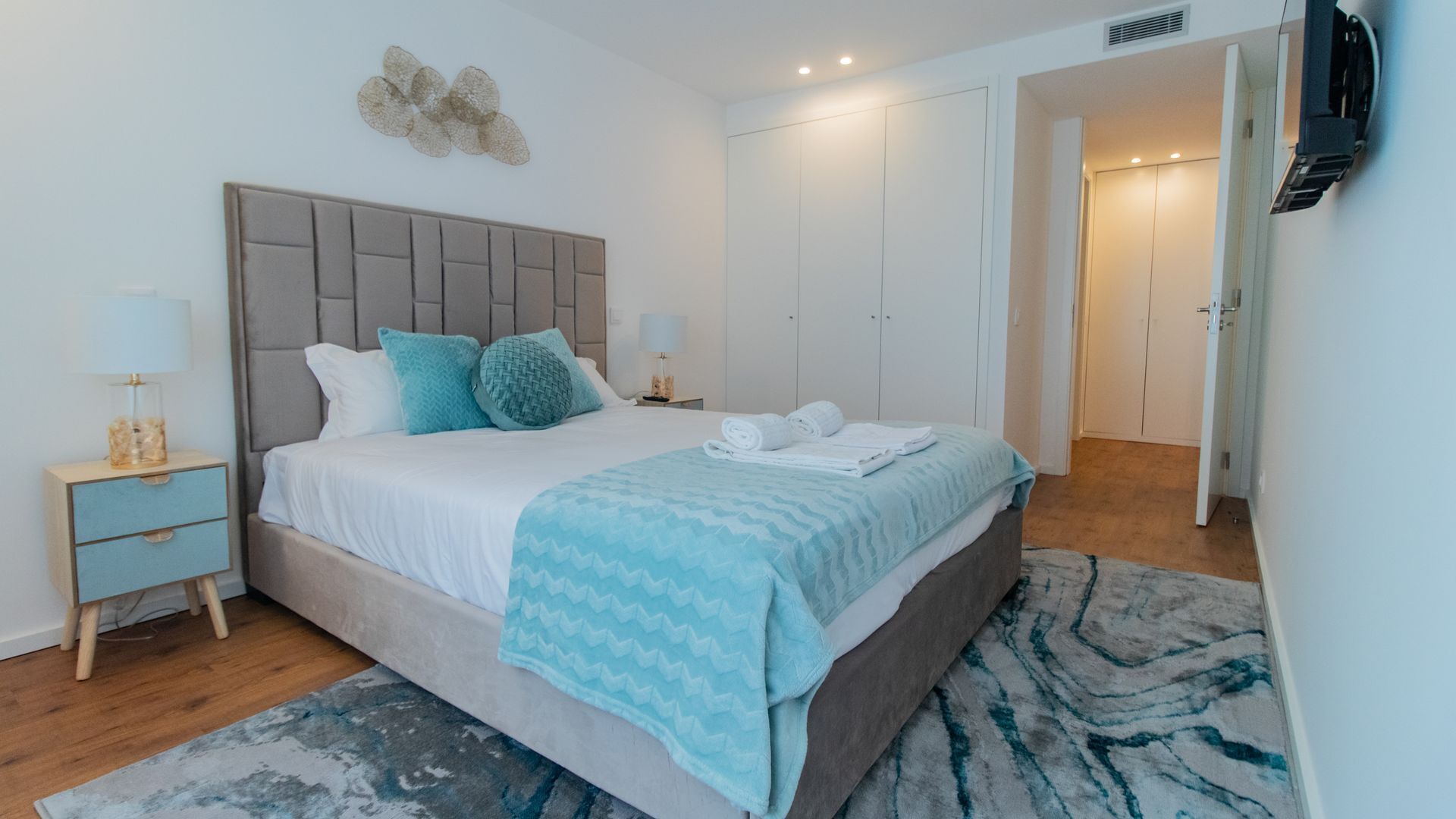 Een moderne slaapkamer met een bed, nachtkastjes, een tv aan de muur en een deur naar een andere kamer.