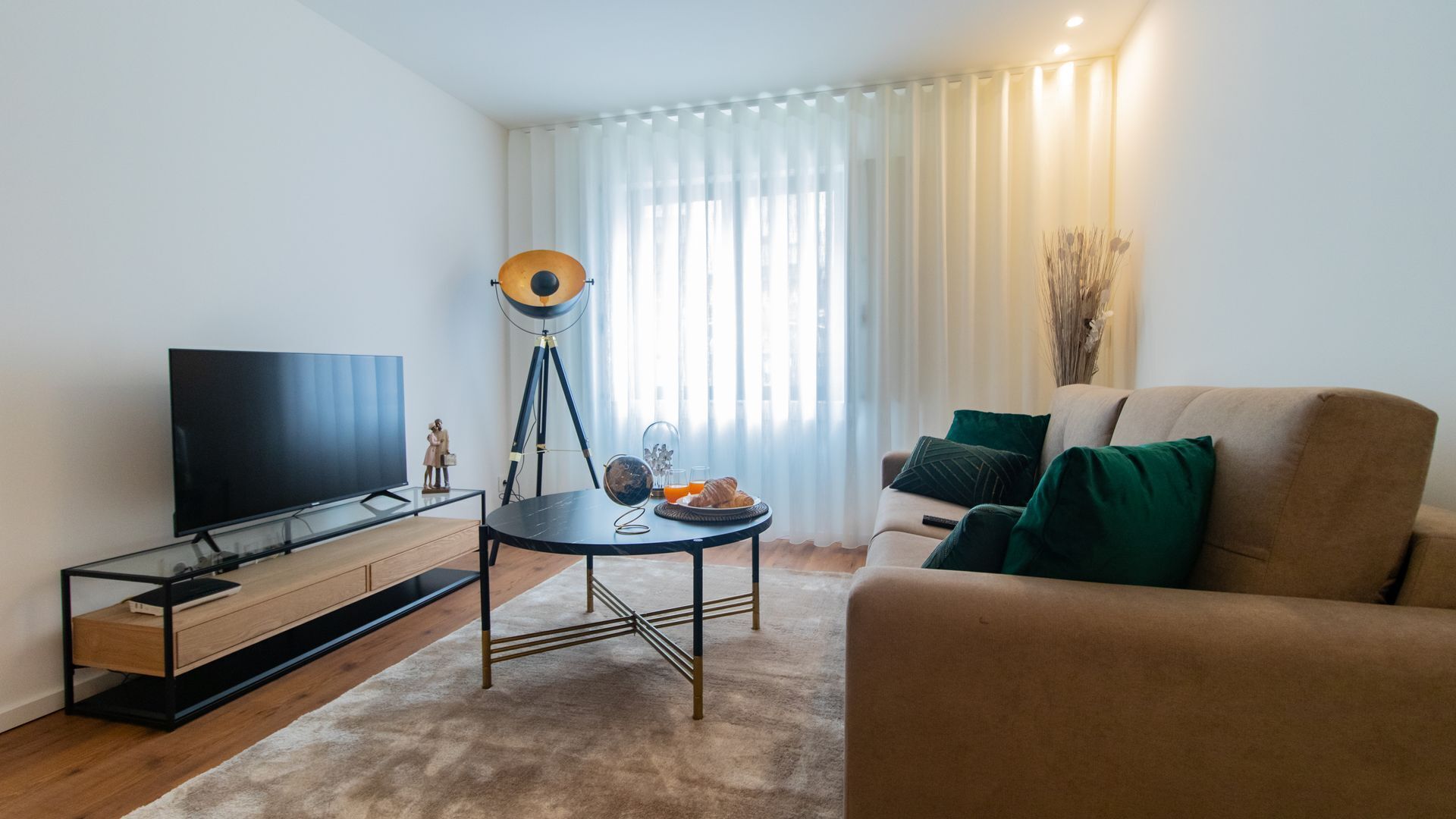 A modern living room with a flat-screen TV, a tripod with a camera, a round table with breakfast items, and a sofa with green cushions.