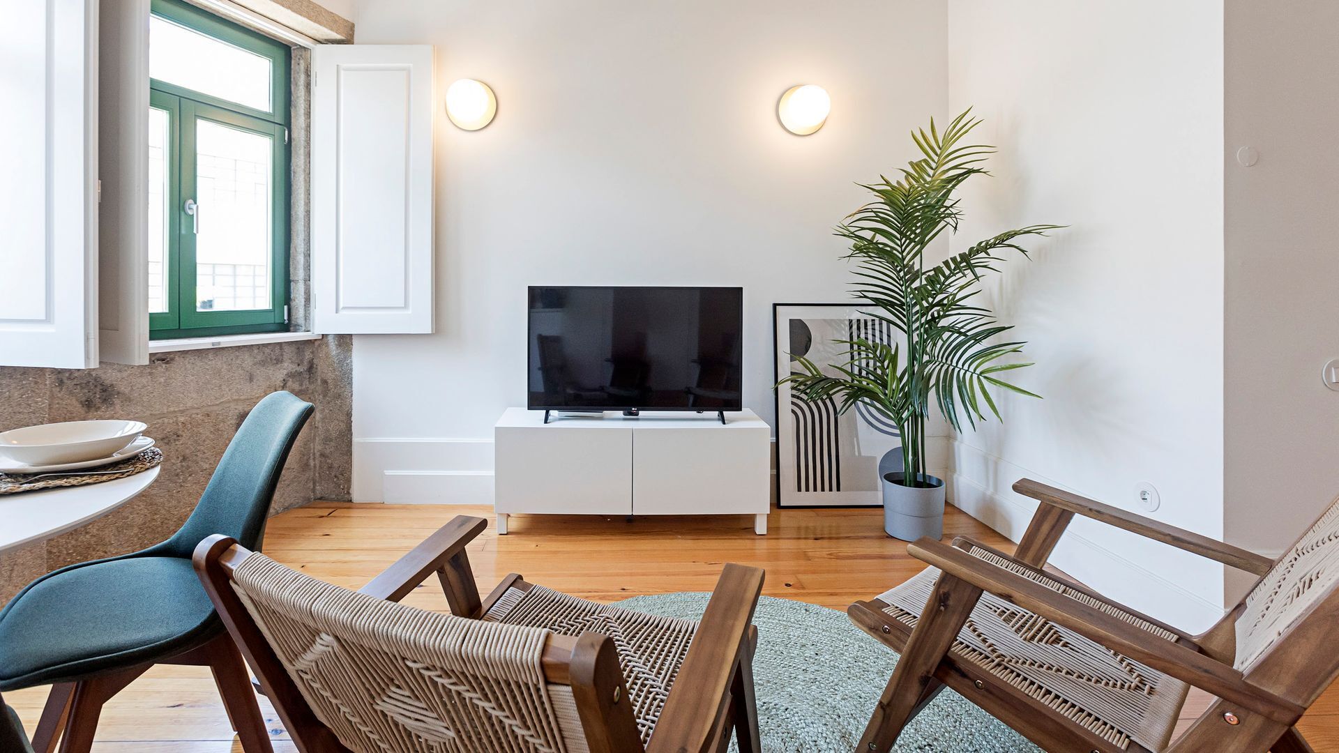 A modern living room with a flat-screen TV on a white cabinet, a dining table with chairs, a potted plant, and a window with green shutters.