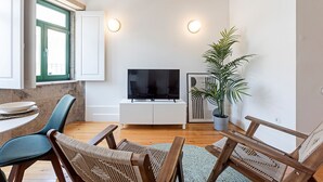 Moderni olohuone, jossa on valkoinen TV-taso ja siinä litteänäyttöinen televisio, ruokapöytä tuoleineen, ruukkukasvi ja ikkuna, jossa on vihreät sälekaihtimet.