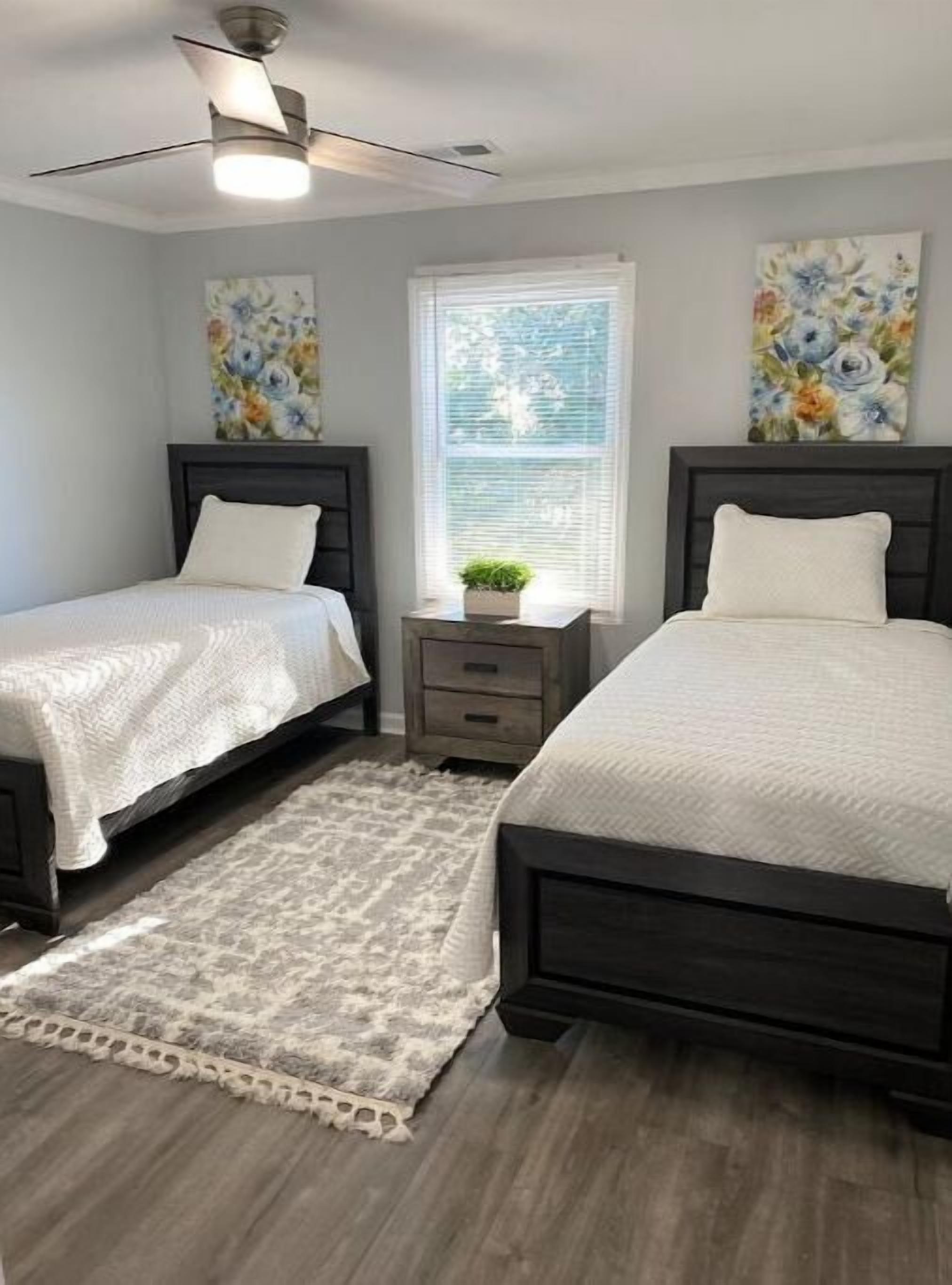 A bedroom with two beds, a ceiling fan, a window with blinds, and floral artwork on the walls.