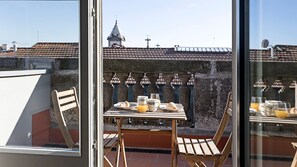 Varanda com mesa posta para o café da manhã, incluindo croissants e suco de laranja, com vista para uma igreja com telhado de azulejos e parede de pedra.