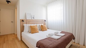 Cama bem arrumada com lençóis brancos e duas almofadas decorativas, cabeceira de madeira e janela com cortinas leves.