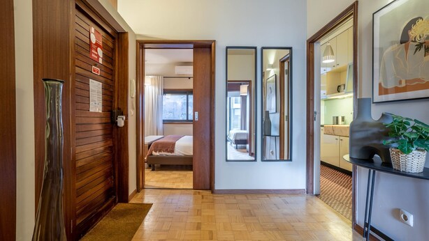 Een hotelgang met een houten deur, een bed in de aangrenzende kamer, een spiegel en een plant op een plank.