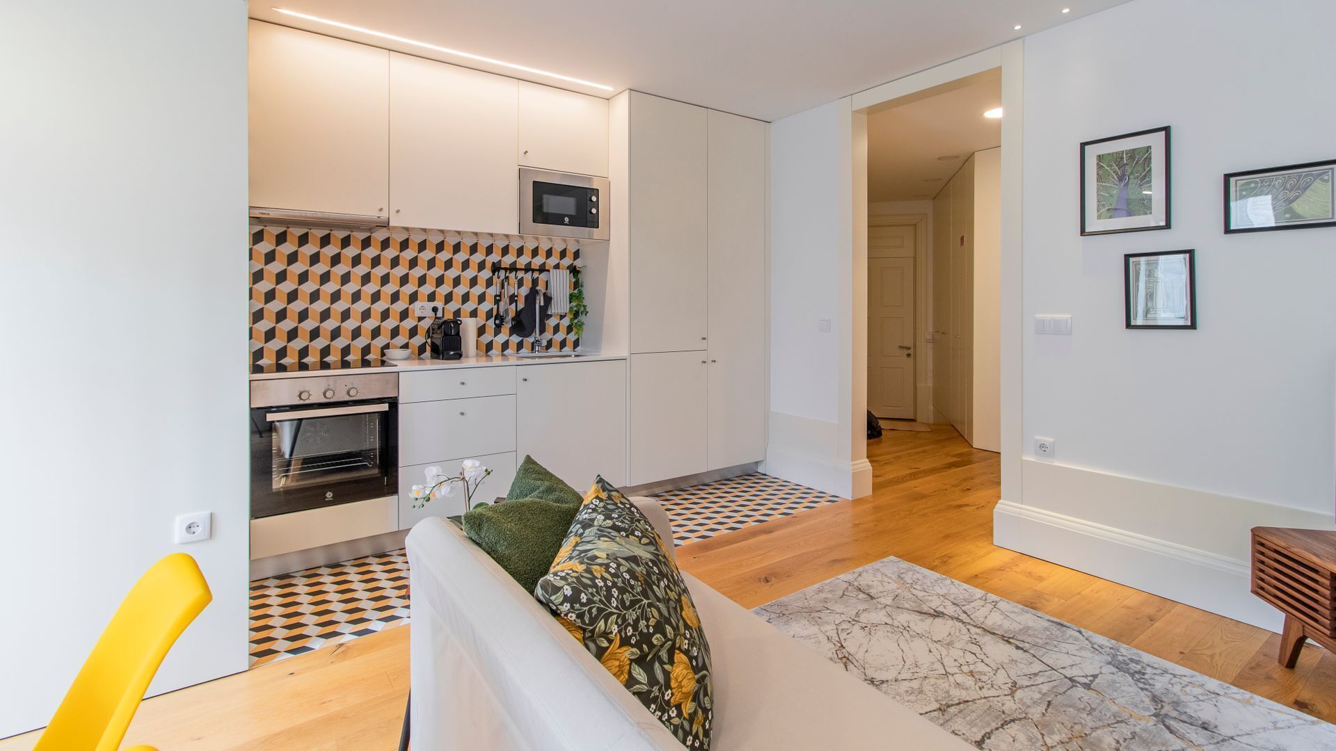 A compact kitchen with white cabinets, a microwave, and an oven. There is a patterned rug and a sofa with cushions.