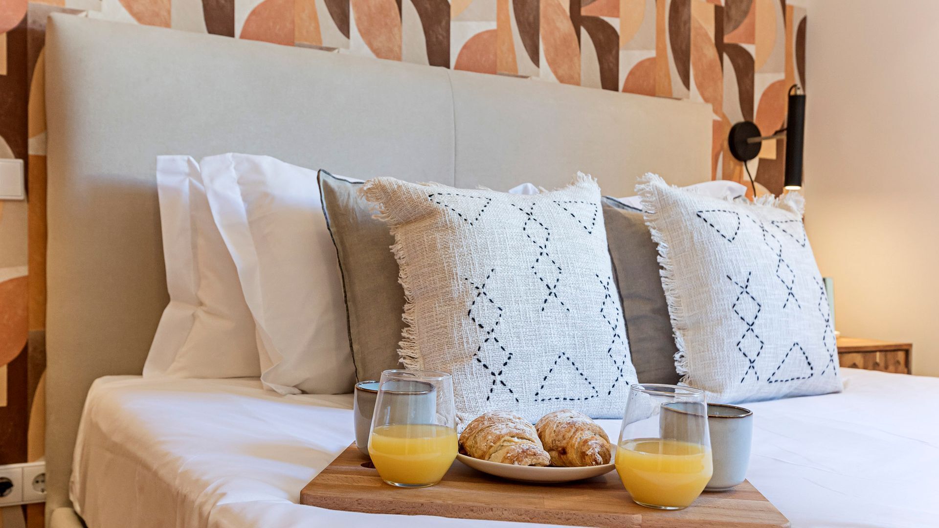 Un lit bien fait, avec du linge de lit blanc, deux oreillers, un plateau avec des croissants et du jus d’orange, et une table de chevet avec une tasse.
