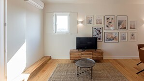 Ein modernes Wohnzimmer mit Flachbildfernseher auf einem Holzschrank, einem Essbereich mit rundem Tisch und einem großen Fenster, das für natürliches Licht sorgt.