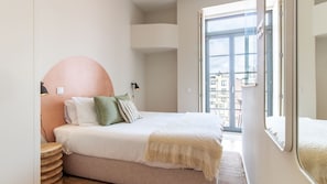 A bedroom with a bed, pillows, a bedside lamp, a round mirror, and a view of buildings through a window.