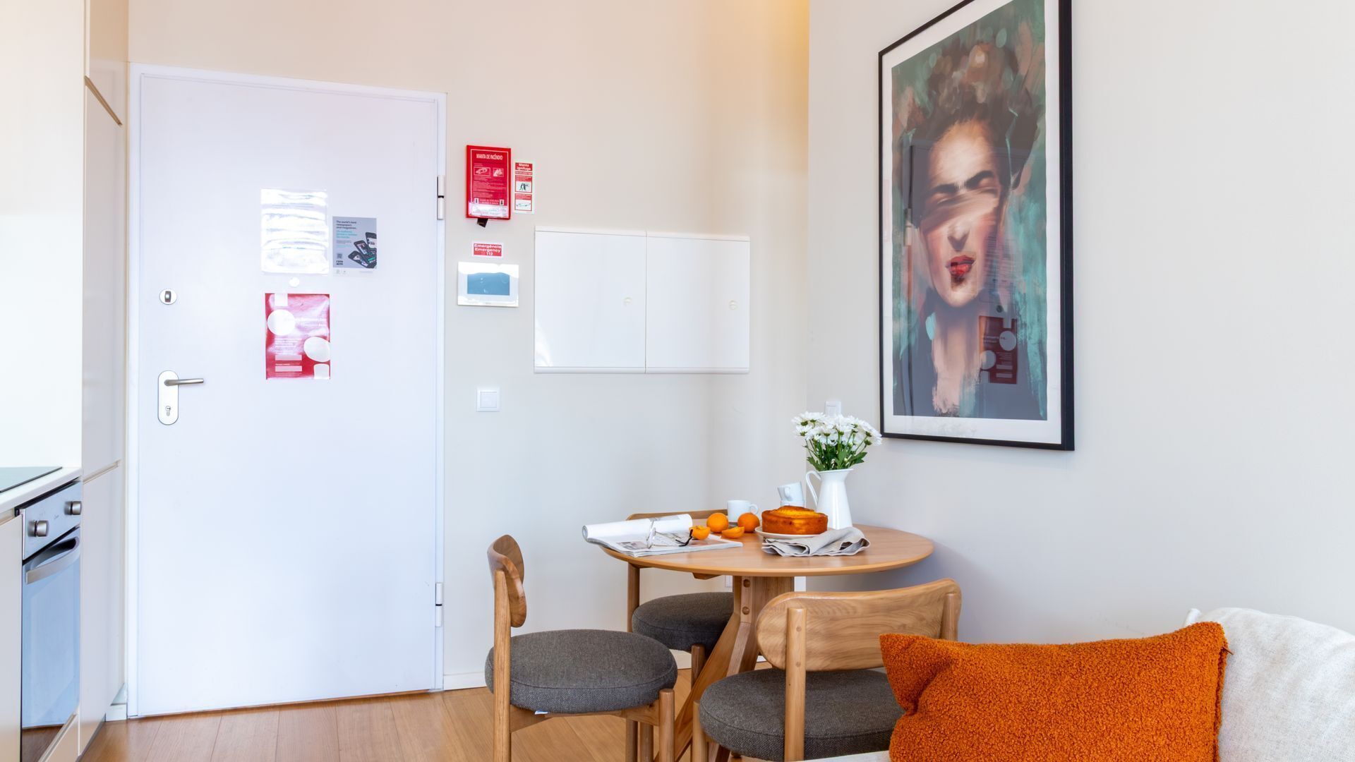 A small, well-lit room with a dining table, chairs, a white door, and a framed picture on the wall.