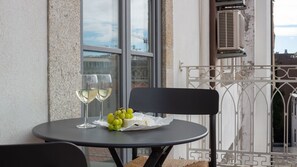 Une table avec deux verres à vin et un panier de raisins, placée devant la façade d’un immeuble doté de grandes fenêtres et d’une rambarde en métal.