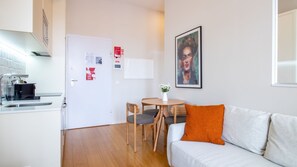 Une cuisine moderne équipée d’une machine à café, une porte blanche ornée d’avis, un coin repas avec une table et des chaises, et un salon avec un canapé et un tableau.