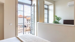Une chambre d’hôtel avec un lit, une fenêtre donnant sur des immeubles, une plante en pot et un climatiseur fixé au mur.