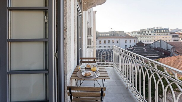 Un balcón con una mesa lista para el desayuno, incluyendo croissants y zumo de naranja, con vistas a una ciudad y edificios tradicionales.