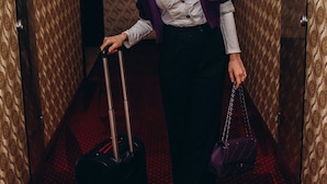 A person in a hallway with patterned wallpaper and a red carpet, holding a suitcase and a purple handbag. - Hotel Novera (Timisoara)