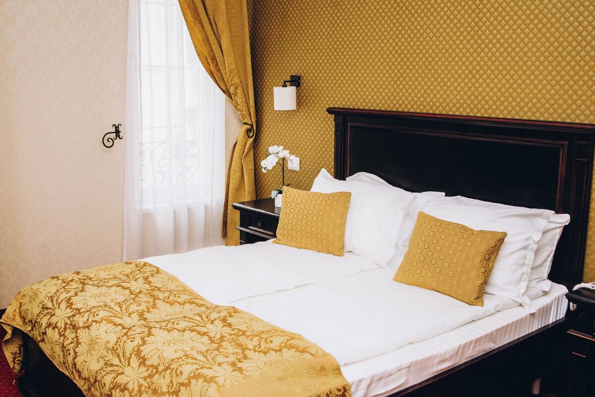 Uma cama bem arrumada com colcha estampada e dois travesseiros, uma mesa de cabeceira com abajur e uma janela com cortinas.