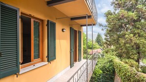 A yellow building with green shutters, a balcony with a metal railing, and a view of trees and a red-roofed building in the background. - Belvilla by OYO Casa Cristina Meina Centro 4 (Meina)