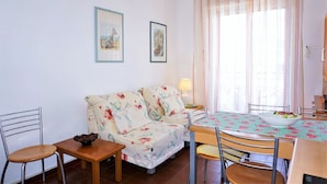 A cozy living room with a sofa, a dining table, and chairs. There are two framed pictures on the wall, a lamp on the table, and a window with curtains. - Holiday Home Caorle Near Beach (Caorle)