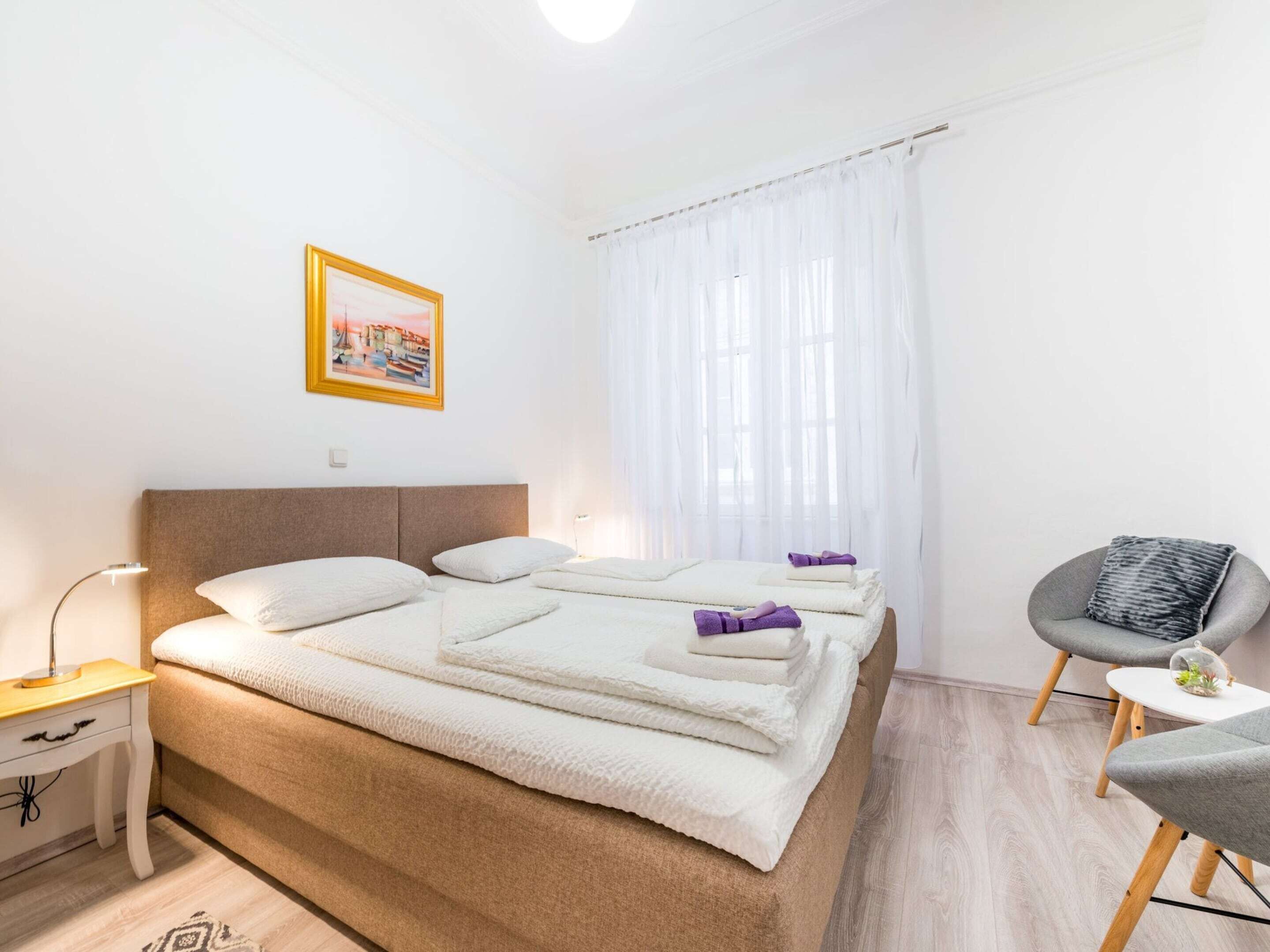 A modern hotel room with a large bed, a bedside table, a chair, and a window with sheer curtains.