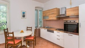 A compact kitchen with white cabinets, a gas stove, and a microwave. There is a dining table with chairs and a window with blinds. - Apartment Tranquilo- Two Bedroom Apartment With Garden View (Dubrovnik)