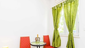 A small room with a table and two chairs, a window with green patterned curtains, and a yellow lamp on a wooden table. - Apartments Versus- Studio Apartment With Terrace (Zupa dubrovacka)