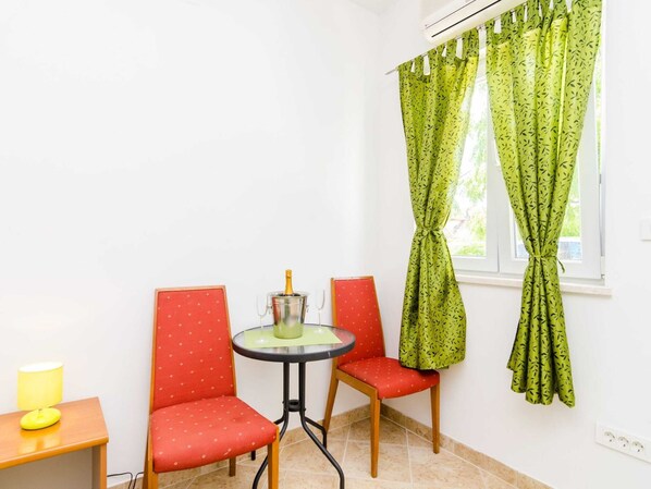 A small room with a table and two chairs, a window with green patterned curtains, and a yellow lamp on a wooden table.