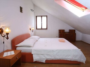A bedroom with a sloped ceiling, a bed with white linens, two wall-mounted lamps, a window with lace curtains, and a wooden dresser. - Apartments Boro - Two Bedroom Apartment With Sea View (Supetar)
