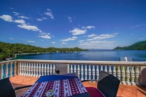 A terrace with a table and chairs, overlooking a calm sea and green hills.